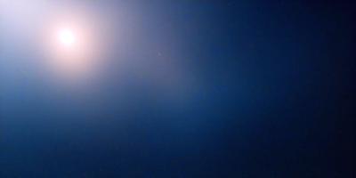 The primary subject of the image is a bright star in the night sky. The setting is a dark, cloudy night with a blue background. The visual style is a photo of the night sky, capturing the star's light. The colors and mood of the image are predominantly dark and moody, with the star being the main source of light in the scene.