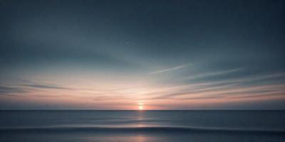 The primary subject of the image is the sunset, with the sun setting over the ocean. The setting is a vast body of water, and the visual style is a photo. The colors and mood of the image are dominated by the warm hues of the sunset, creating a serene and peaceful atmosphere.