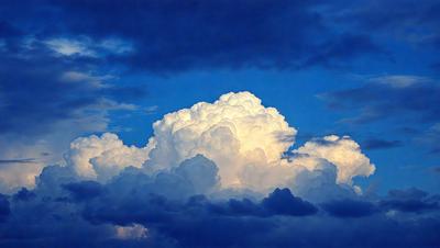The primary subject of the image is a large, fluffy cloud in the sky. The cloud is surrounded by a blue sky, creating a serene and peaceful atmosphere. The setting is outdoors, with the cloud as the main focus of the scene. The visual style of the image is a photo, capturing the beauty of the cloud in its natural environment. The colors in the image are predominantly blue and white, with the cloud being predominantly white, and the sky being a deep shade of blue.