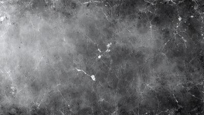 The primary subject of the image is a gray, textured surface, which appears to be a wall or a piece of art. The setting is a dark room, and the visual style is a close-up of the textured surface. The colors and mood of the image are predominantly dark and moody, with the gray surface being the main focus.