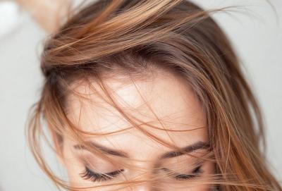 The primary subject of the image is a woman with blonde hair. She has her eyes closed, and her hair is blowing in the wind. The setting is outdoors, with the woman standing in front of a white background. The visual style is a close-up of the woman's face, and the colors are predominantly shades of brown and white. The overall atmosphere is serene and peaceful.