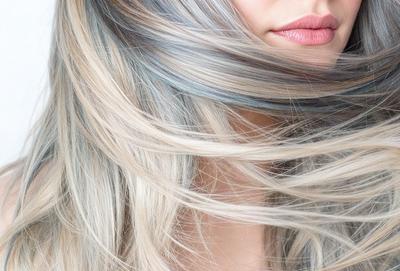 The primary subject of the image is a woman with blonde hair. She is blowing her hair back and has a blue streak in it. The setting is outdoors, with the woman standing in front of a blue sky. The visual style is a photo, capturing the woman's natural beauty. The colors and mood of the image are bright and cheerful, with the blue streak adding a touch of color and personality to the woman's hair.