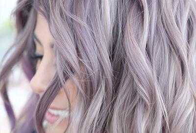 The primary subject of the image is a woman with purple hair. She is smiling and has her hair blowing in the wind. The setting is outdoors, with the woman standing in front of a building. The visual style is a photo, capturing the woman's unique appearance and the natural lighting. The colors and mood of the image are vibrant and lively, with the purple hair adding a touch of whimsy to the scene.