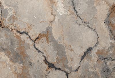 The primary subject of the image is a cracked and chipped stone wall. The setting is a stone wall with a rusty appearance, giving it a worn and aged look. The visual style is a close-up of the stone wall, showcasing its texture and details. The colors and mood of the image are predominantly brown and grey, with a sense of decay and weathering.