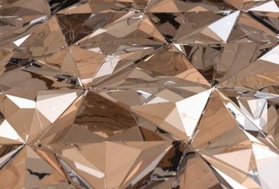 The primary subject of the image is a close-up of a shiny, reflective surface, possibly a mirror or a piece of artwork. The setting is a room with a white background, which provides a clean and minimalistic environment. The visual style appears to be a combination of a photo and a digital artwork, as the surface appears to be both reflective and textured. The colors in the image are predominantly silver and gold, creating a warm and elegant atmosphere.