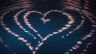 The primary subject of the image is a heart-shaped pattern created by ripples in the water. The heart is made up of multiple smaller hearts, which are the ripples in the water. The setting is a body of water, and the visual style is a close-up of the heart-shaped pattern. The colors and mood of the image are predominantly blue, with a calm and serene atmosphere.