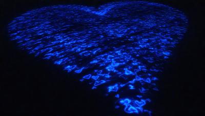 The primary subject of the image is a blue heart-shaped pattern, which is displayed on a blue surface. The setting appears to be a dark room, and the visual style is a combination of a photo and digital art. The dominant colors in the image are blue and purple, creating a calm and serene atmosphere.