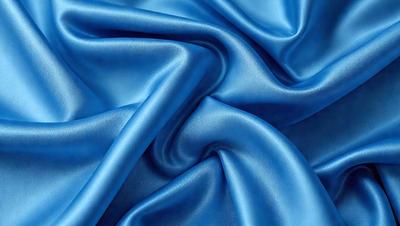 The primary subject of the image is a blue fabric, which appears to be a piece of silk. The setting is a close-up view of the fabric, showcasing its texture and color. The visual style is a realistic representation of the fabric, with no indication of digital art or illustration. The dominant colors are blue and silver, creating a calm and soothing atmosphere.