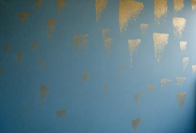 The primary subject of the image is a wall with a blue background. The wall is decorated with a pattern of gold stars, creating a visually appealing and unique design. The setting appears to be a room with a blue color scheme, which complements the gold stars on the wall. The visual style of the image is a combination of artistic elements, such as the gold stars and the blue background, creating a distinctive and eye-catching design. The colors and mood of the image are predominantly blue and gold,