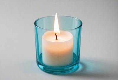 The primary subject of the image is a lit candle in a glass cup. The candle is placed in the center of the cup, and the glass cup is filled with water. The setting is a table, and the visual style is a close-up of the candle in the glass cup. The colors and mood of the image are warm and inviting, with the lit candle creating a cozy atmosphere.