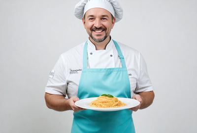 The primary subject of the image is a man wearing a chef's outfit, holding a plate of spaghetti. The man is smiling, and the plate of spaghetti is placed in front of him. The setting appears to be a kitchen or a dining area, as the man is dressed in a chef's uniform and is holding a plate of food. The visual style of the image is a photo, and the colors and mood are warm and inviting, with the man's smile adding a sense of