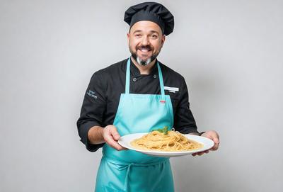 The primary subject of the image is a man wearing a chef's outfit, holding a plate of spaghetti. He is smiling and appears to be proud of his culinary creation. The setting is a kitchen, with a dining table visible in the background. The visual style is a photo, capturing the man and his dish in detail. The colors and mood of the image are warm and inviting, with the man's smile and the delicious-looking spaghetti creating a sense of enjoyment