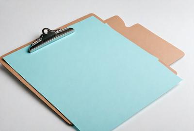 The primary subject of the image is a clipboard with a blue cover. The clipboard is placed on a white surface, which serves as the setting or background. The visual style of the image is a photo, capturing the details of the clipboard and its surroundings. The dominant colors in the image are blue and white, creating a calm and organized atmosphere.