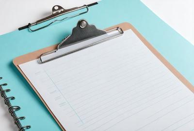 The primary subject of the image is a clipboard with a piece of paper on it. The paper is white and has writing on it. The clipboard is attached to a metal clip, which is placed on a table. The setting is an indoor environment, and the visual style is a photo. The colors and mood of the image are predominantly white and neutral, giving it a clean and organized appearance.