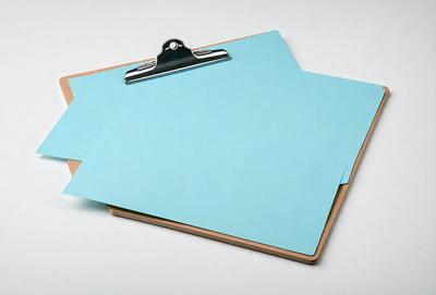 The primary subject of the image is a blue clipboard with a metal clip. The clipboard is placed on a white surface, which serves as the setting or background. The visual style of the image is a photo, capturing the details of the clipboard and the metal clip. The dominant colors in the image are blue and white, creating a calm and organized atmosphere.