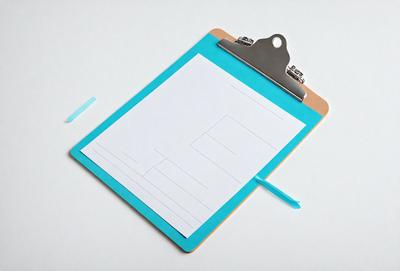 The primary subject of the image is a clipboard with a blue cover. The clipboard is placed on a white surface, which serves as the setting or background. The visual style of the image is a photo, capturing the details of the clipboard and its surroundings. The dominant colors in the image are blue and white, creating a clean and organized appearance.