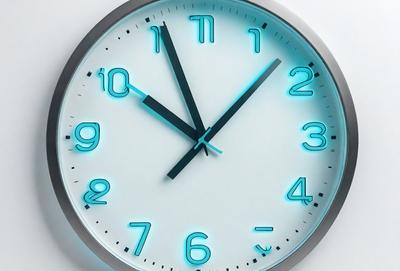 The primary subject of the image is a clock with a blue face and black hands. The clock is set against a white background, giving it a clean and modern appearance. The visual style of the image is a photo, capturing the details of the clock's design and the contrast between the blue face and the white background. The colors and mood of the image are predominantly blue and white, creating a sense of calm and orderliness.