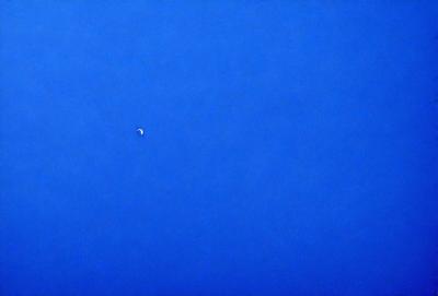 The primary subject of the image is the moon, which is visible in the sky. The setting is a clear blue sky, and the visual style is a photo. The colors and mood of the image are predominantly blue, creating a calm and serene atmosphere.