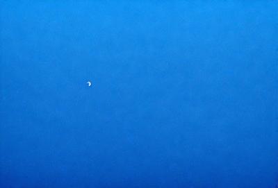 The primary subject of the image is the moon, which is visible in the sky. The setting is a clear blue sky, with no clouds or other obstructions. The visual style is a photo, capturing the moon in its natural environment. The colors and mood of the image are predominantly blue, creating a serene and peaceful atmosphere.