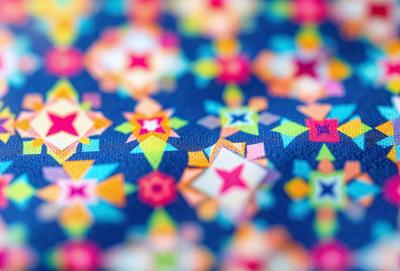 The primary subject of the image is a colorful, patterned fabric with a blue background. The fabric features a variety of stars and other shapes, creating a visually appealing and vibrant design. The setting appears to be a close-up of the fabric, showcasing its intricate details. The visual style of the image is a combination of digital art and illustration, as the fabric's design is both complex and well-defined. The colors and mood of the image are lively and cheerful, with the