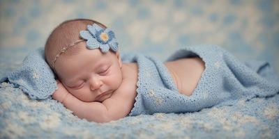 The primary subject of the image is a baby girl wearing a blue knit cap with a blue flower on it. She is laying down on a blue blanket, which is placed on a blue surface. The visual style of the image is a photo, capturing the baby's peaceful and innocent expression. The colors and mood of the image are predominantly blue, creating a calm and soothing atmosphere.