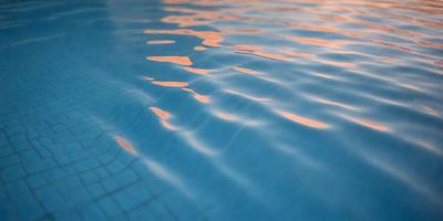The primary subject of the image is a swimming pool filled with water. The pool is blue and has a few ripples on the surface. The setting is an outdoor environment, and the visual style appears to be a photo. The colors and mood of the image are calm and serene, with the blue water and ripples creating a peaceful atmosphere.