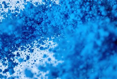 The primary subject of the image is a blue substance, possibly a liquid or a powder, that is dispersed throughout the scene. The setting appears to be a white background, which contrasts with the blue substance. The visual style of the image is a close-up of the blue substance, showcasing its texture and appearance. The dominant colors in the image are blue and white, creating a calm and soothing atmosphere.