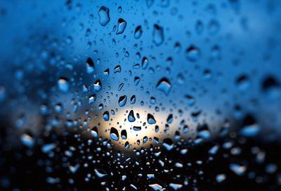 The primary subject of the image is a rainy day, as seen through a window. The setting is an outdoor environment, with a cloudy sky visible in the background. The visual style is a photo, capturing the raindrops on the window and the cloudy sky. The colors and mood of the image are predominantly dark and moody, with the raindrops and cloudy sky creating a sense of gloominess.