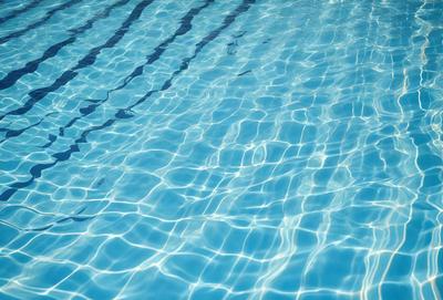 The primary subject of the image is a swimming pool filled with water. The pool is blue and has a clear, reflective surface. The setting is an outdoor environment, with the pool being the main focus of the scene. The visual style of the image is a photo, capturing the natural light and reflections on the water's surface. The colors in the image are predominantly blue, creating a calm and relaxing atmosphere.