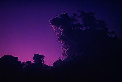 The image features a purple sky with a cloudy atmosphere. The primary subject is a large cloud that appears to be forming a face, giving the scene a unique and artistic visual style. The setting is a nighttime sky, and the colors and mood convey a sense of mystery and intrigue.