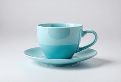 The primary subject of the image is a blue coffee cup with a handle, placed on a white surface. The cup is positioned on a small plate, which is resting on a larger plate. The setting appears to be a simple and clean environment, with no additional objects or distractions. The visual style of the image is a close-up of the coffee cup, emphasizing its color and details. The dominant colors in the image are blue and white, creating a calm and soothing atmosphere.