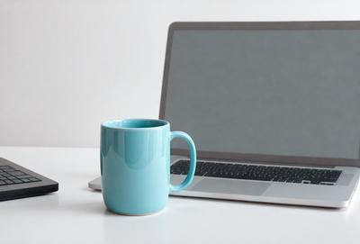 The primary subject of the image is a laptop computer. The laptop is placed on a white surface, which serves as the setting for the scene. The visual style of the image is a photo, capturing the laptop and the cup of coffee placed next to it. The dominant colors in the image are white and blue, creating a clean and calming atmosphere.