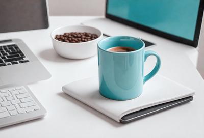 The primary subject of the image is a blue coffee mug placed on a white desk. The setting is a workspace, with a laptop and a bowl of coffee beans nearby. The visual style is a photo, and the colors and mood are predominantly white and blue, creating a calm and focused atmosphere.