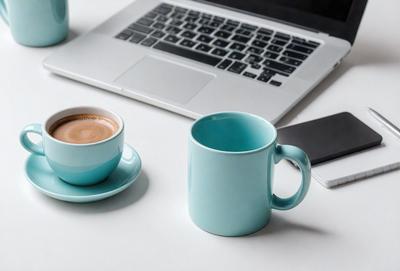 The primary subject of the image is a laptop computer, which is placed on a white surface. The laptop is accompanied by two cups of coffee, one of which is a blue mug. The setting appears to be a workspace or a home office, with the laptop and coffee cups as the main focus. The visual style of the image is a realistic representation of the laptop and coffee cups, with no indication of digital art or illustration. The colors and mood of the image are predominantly neutral, with the
