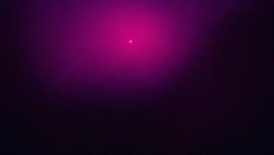 The primary subject of the image is a bright pink circle, which appears to be a sun or a light source. The setting is a dark, purple background, creating a contrast between the pink circle and the surrounding area. The visual style is a combination of a photo and a digital art, as the pink circle stands out against the dark background. The colors and mood of the image are predominantly dark and moody, with the pink circle being the only bright and vibrant element in the scene.