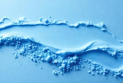 The primary subject of the image is a blue substance, possibly a liquid or a powder, that is spreading or dripping onto a white surface. The setting appears to be a table or a countertop, with the blue substance creating a visually striking contrast against the white background. The visual style of the image is a close-up, focusing on the details of the blue substance and its interaction with the white surface. The colors and mood of the image are predominantly blue and white, creating a sense of