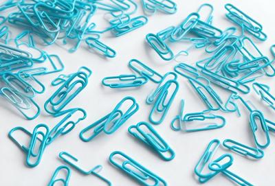 The primary subject of the image is a collection of blue paper clips. The setting is a white surface, which serves as a backdrop for the clips. The visual style is a close-up of the paper clips, showcasing their vibrant blue color. The overall atmosphere is organized and clean, with the blue paper clips neatly arranged on the white surface.