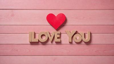 The primary subject of the image is a wooden heart with the words "I Love You" written on it. The heart is placed on a pink background, which adds a warm and romantic touch to the scene. The visual style of the image is a combination of a photo and digital art, creating a unique and personalized display of love. The colors and mood of the image are predominantly pink, which is often associated with love and affection.