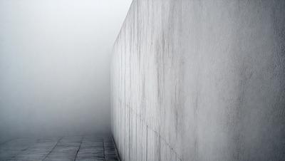The primary subject of the image is a tall, white, concrete wall. The setting is a foggy, misty environment, creating a mysterious and atmospheric mood. The visual style is a photo, capturing the intricate details of the concrete wall. The colors and mood are predominantly gray and moody, with the fog and mist adding a sense of depth and enigma to the scene.
