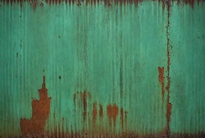 The primary subject of the image is a green metal surface with rust and dirt on it. The setting is an outdoor environment, possibly a building or a structure. The visual style is a close-up of the green metal surface, which appears to be a rusted metal wall. The colors and mood of the image are dominated by the green color of the metal and the rust, giving it a somewhat aged and weathered appearance.