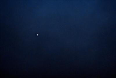 The primary subject of the image is a moon, which is visible in the sky. The setting is a dark, cloudy night with a blue sky. The visual style is a photo, capturing the moon and the night sky. The colors and mood of the image are predominantly dark and moody, with the moon being the main source of light in the scene.