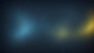 The primary subject of the image is a yellow light, which is the main focus of the scene. The setting is a dark, foggy environment, with a blue background. The visual style appears to be a photo, as the light is captured in a clear and detailed manner. The colors and mood of the image are predominantly dark and moody, with the yellow light providing a contrasting and warm element.