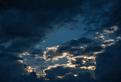 The primary subject of the image is a cloudy sky with a sunset in the background. The setting is a dark, cloudy sky, and the visual style is a photo. The colors and mood of the image are dominated by the warm hues of the sunset and the dark, moody clouds.