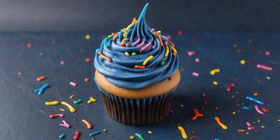 The primary subject of the image is a cupcake with a blue frosting and colorful sprinkles on top. The cupcake is placed on a black surface, which serves as the setting or background. The visual style of the image is a close-up of the cupcake, showcasing its details and colors. The dominant colors in the image are blue, brown, and various shades of colorful sprinkles. The overall atmosphere of the image is cheerful and inviting, as the cupcake
