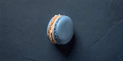 The primary subject of the image is a small, round, blue and white pastry, which appears to be a macaroon. The pastry is placed on a blue surface, which could be a table or a counter. The visual style of the image is a close-up, focusing on the details of the pastry. The colors and mood of the image are predominantly blue and white, giving it a clean and fresh appearance.