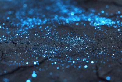 The primary subject of the image is a blue light, which is shining brightly on a dark surface. The setting appears to be a dirt ground, and the visual style is a close-up of the blue light source. The colors and mood of the image are predominantly dark and moody, with the blue light providing a contrasting and captivating element.