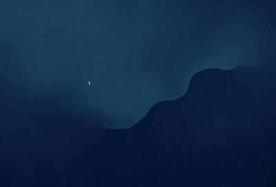 The primary subject of the image is a moon, which is seen in the sky. The setting is a dark, cloudy night, with the moon being the main focus of the scene. The visual style is a combination of a photo and digital art, creating a unique and artistic representation of the moon. The colors and mood of the image are predominantly dark and moody, with the moon being the only source of light in the scene.