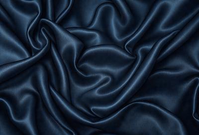 The primary subject of the image is a close-up of a blue fabric, possibly a piece of silk. The setting is a dark background, which emphasizes the richness of the blue fabric. The visual style is a close-up, which highlights the intricate details of the fabric. The colors and mood of the image are predominantly blue, creating a sense of calmness and sophistication.