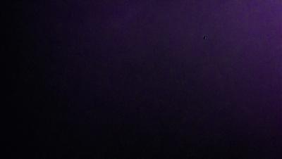 The image is a black and white photo of a purple background. The primary subject is a small dot, which is located towards the top right corner of the image. The setting is a dark, purple background, and the visual style is a black and white photo. The colors and mood of the image are predominantly purple, creating a moody and dramatic atmosphere.