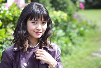 The primary subject of the image is a woman wearing a purple jacket and a pearl necklace. She is posing for the camera and smiling. The setting is outdoors, with a garden in the background. The visual style is a photo, and the colors and mood are warm and inviting.